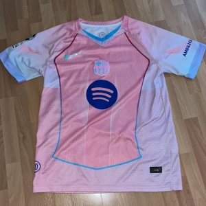 Barça Lamine Yamal rosa matchtröja - Säljer en Barcelona fotbollströja med Lamine Yamal och nummer 10 på ryggen. Tröjan är rosa med ljusblå och vita detaljer, har korta ärmar och är tillverkad i lätt, ventilerande polyester. Framsidan har Spotify-logga och klubbmärke. 