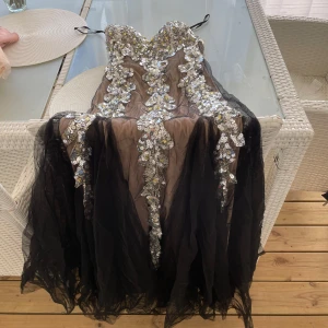 Svart långklänning med paljetter - Säljer en galet snygg svart långklänning med transparent mesh och glittriga silver- och guldfärgade paljetter framtill och baktill. Klänningen har en axelbandslös design och snörning i ryggen för extra cool passform. Perfekt för dig som vill sticka ut på bal eller fest!