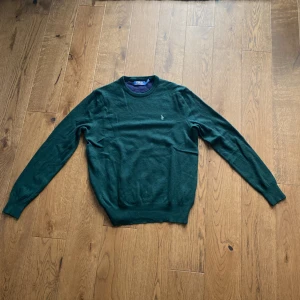 Mörkgrön stickad tröja Polo Ralph Lauren - Snygg mörkgrön stickad tröja från Polo Ralph Lauren med klassisk rund hals och broderad logga på bröstet. Tröjan har långa ärmar och ribbade muddar vid hals, ärmslut och nederkant. Perfekt för dig som gillar stilrena och tidlösa plagg.