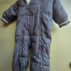 Mörkgrå pufferoverall från KappAhl - Mysig mörkgrå pufferoverall från KappAhl med två dragkedjor framtill för enkel påklädning. Overallen har huva, ribbade muddar med vit rand och är fodrad med mjuk fleece på insidan. Perfekt för kalla dagar när du vill hålla dig varm och bekväm.