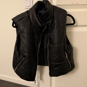 Svart dunväst i skinn med dragkedja - Snygg svart skinnväst med hög krage och två snedställda fickor med dragkedjor. Västen har quiltad design och en kraftig dragkedja framtill. Perfekt för lager-på-lager och ger en cool vibe till din outfit.