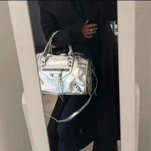 Silver handväska  - Säljer en snygg handväska  i metallic silver. Väskan har klassiska detaljer med dragkedja framtill, handtag och en axelrem. Perfekt storlek för att få plats med allt du behöver och ger en cool vibe till din outfit.