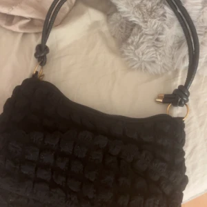 Svart quiltad axelväska med guld-detaljer - Snygg svart axelväska med quiltad yta och mjukt material. Väskan har två rejäla handtag med guldfärgade ringar och dragkedja upptill. Insidan är ljus och har en praktisk ficka för småsaker. Perfekt storlek för det viktigaste.