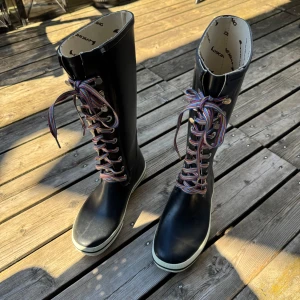 Svarta Viking snörade stövlar - Svarta höga stövlar från Viking med färgglada randiga skosnören och vit kant runt sulan. Stövlarna har rund tå, platt sula och är tillverkade i syntetmaterial som tål regn. Perfekt för dig som vill sticka ut med coola detaljer och färg på snörningen.