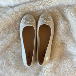 Vita ballerinaskor med rosett - Snygga vita ballerinaskor från Fashion med rund tå och dekorativa rosetter framtill. Skorna har en glansig finish och är tillverkade i syntetmaterial. Platt sula och klassisk design som passar till många olika outfits. Perfekt för dig som gillar stilrena och söta detaljer.