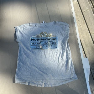 Grå t-shirt med paljetter Marc Jacobs - Snygg grå t-shirt från Little Marc Jacobs med coolt paljettmotiv i silver och guld framtill. T-shirten har korta ärmar och ett statement-tryck med texten 'Now Showing Marc Jacobs Spring 2017'. Mjuk bomullskvalitet och relaxed fit. Ny pris ligger på 890kr