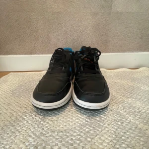 Svarta Adidas sneakers med blå detaljer - Säljer ett par svarta Adidas sneakers med blå ränder och orange logga på hälen. Skorna har snörning, vit mellansula och svart yttersula med blå inslag. Insidan är fodrad med blått tyg och materialet är syntet och skinn. Perfekt för dig som gillar sportig stil och färgkombinationer.