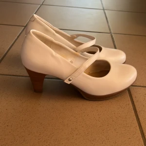 Vita pumps med klack från Gyllene Gripen - Snygga vita pumps från Gyllene Gripen med rund tå och klassisk Mary Jane-rem över foten. Skorna har en träfärgad klack i medelhöjd och är tillverkade i slätt syntetmaterial. Perfekt för dig som gillar stilrena och feminina detaljer.