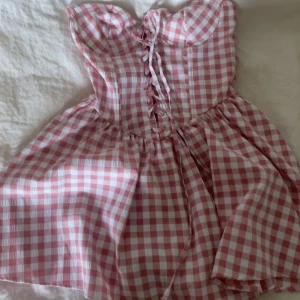 Rutig rosa kortklänning med snörning - Supersöt kortklänning i rosa och vitt rutigt mönster. Klänningen är axelbandslös och har en snygg snörning framtill som ger en cool touch. Den har en utställd kjol och är gjord i ett lätt bomullsmaterial, perfekt för varma dagar. Passformen är normal och stilen är riktigt trendig.