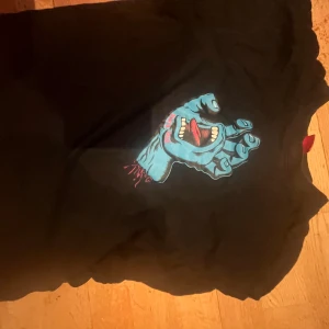 Svart Santa Cruz t-shirt med tryck - Svart t-shirt från Santa Cruz i storlek M med ikoniskt blått handtryck på bröstet, där handen har en öppen mun med tunga och tänder. T-shirten har rund hals och korta ärmar, perfekt för dig som gillar streetwear och skatekultur.