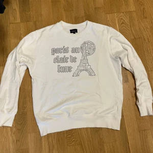 Vit sweatshirt med Rhinestones - Vit sweatshirt  med tryck i Rhinestones som föreställer Eiffeltornet och texten 'paris au clair de lune' på framsidan. Tröjan har rund hals, långa ärmar och ribbade muddar vid ärmslut och nederkant. Perfekt för dig som gillar Paris vibes. Storlek L, passar M. Några fåtal stenar har lossnat.
