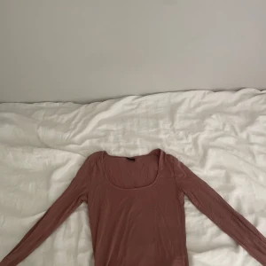 Rosa långärmad topp med fyrkantig hals - Snygg långärmad topp i en mjuk rosa färg med fyrkantig halsringning. Modellen är slim och sitter tajt mot kroppen, perfekt för att skapa en clean och stilren look. Materialet känns lätt och stretchigt, vilket gör den skön att bära hela dagen.