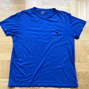 Blå t-shirt från Ralph Lauren - Snygg blå t-shirt från Ralph Lauren med klassisk rund hals och korta ärmar. På bröstet finns en ficka med den ikoniska Polo-loggan broderad i svart. T-shirten är gjord i mjuk bomull och har en normal passform, perfekt för chill dagar.