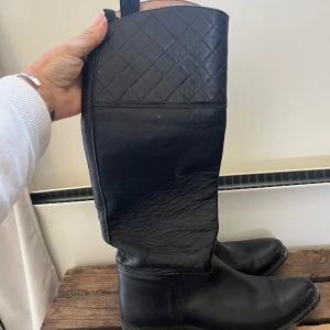 Svarta ridstövlar från El Estribo - Snygga svarta ridstövlar från El Estribo, handgjorda i Spanien. Stövlarna är tillverkade i skinn med quiltad detalj upptill. Perfekta för dig som gillar stilren och tidlös look till ridning.                                                                           Skafthöjd utsida 40cm insida 35cm skaftbredd 34cm 