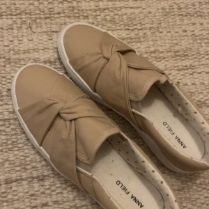 Beige sneakers med rosett från Anna Field - Snygga beige sneakers från Anna Field med en cool rosettdetalj på ovansidan. Skorna har rund tå och platt vit sula, vilket ger en clean och modern look. Insidan är mönstrad med små prickar för extra stilpoäng. Perfekt för dig som gillar enkel men trendig design.