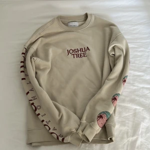 Beige Joshua Tree sweatshirt från Valient - Snygg beige sweatshirt från Valient med tryck 'Joshua Tree' på bröstet och stort grafiskt motiv på ryggen. Ärmarna har små tryck i vinrött och flera runda illustrationer med naturtema i rosa, vinrött och turkos.