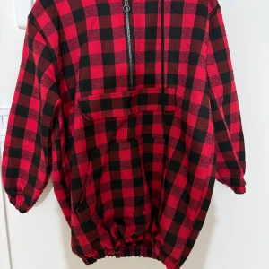 Röd och svart rutig hoodie med huva - Säljer en röd och svart rutig hoodie med huva och half zip framtill. Tröjan har elastisk nederkant och ärmar, stor ficka på magen och ett klassiskt rutmönster. Perfekt för dig som gillar streetstyle och vill sticka ut med en bold look.