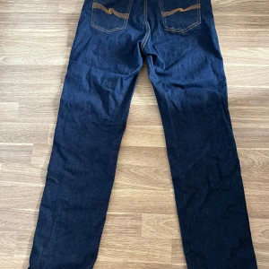 Oanvända Mörkblå jeans från Nudie Jeans, W31 L30 - Oanvända snygga mörkblå jeans från Nudie Jeans med klassisk orange kontrastsöm och raka ben. Modellen har fem fickor och coola detaljer på bakfickorna. Perfekt passform för dig som gillar stilrena och tidlösa jeans. Materialet är slitstarkt denim i bomull.