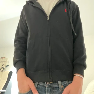 Svart hoodie från Polo Ralph Lauren - Svart hoodie från Polo Ralph Lauren med röd logga broderad på bröstet. Tröjan har huva med snörning, dragkedja framtill och två fickor. Storlek M. Ganska liten k storleken skulle säga att den är mer lik storlek S. Säljer pga av att jag inte använder den! Kan gå ner i pris