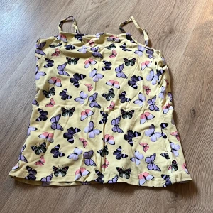 Gul linnetopp med fjärilsmönster - Säljer en söt gul linnetopp med tunna axelband och färgglada fjärilar i lila, rosa och svart över hela toppen. Perfekt för soliga dagar och har en lätt och skön känsla. Den är från Lindex men är köpt för länge sedan. Knappt använd så den är som ny. Storlek 170 men passar xs/s