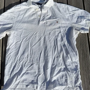 Vit piké t-shirt från GANT - Vit piké t-shirt från GANT med klassisk krage och två knappar vid halsen. Diskret GANT-logga broderad på bröstet. T-shirten är kortärmad och tillverkad i mjuk bomull som andas. Perfekt för dig som gillar stilren och enkel design. Är lätt gulnad i armhålorna (se bild) men är inget märkbart vid användning. Sitter som en tight L