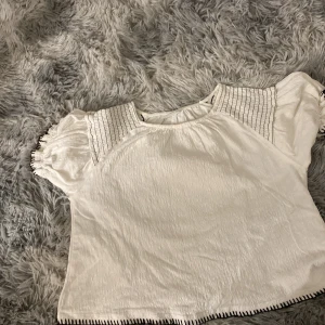 Vit blus med puffärm från Zara - Supersöt vit blus från Zara med puffiga korta ärmar och smockdetaljer vid axlarna. Svarta sömmar längs ärmslut och nederkant ger en cool kontrast. Blusen är gjord i mjukt bomullstyg och har en loose passform som känns chill och bekväm.