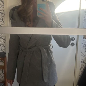 Grå kappa med bälte - Snygg grå kappa från Amisu med bred krage och omlottstängning. Jackan har ett matchande bälte i midjan och knappar framtill. Långärmad och tillverkad i mjukt ullblandat material, perfekt för kyliga dagar. Stilren och enkel att matcha med olika outfits. Nypris 600kr. Den är endast använd några fåtal gånger. Den har en stor luva. Perfekt till kalla höst och vinter dagar!🍁❄️