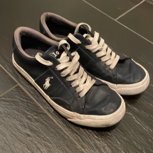 Mörkblå sneakers från Polo Ralph Lauren med Box - Snygga mörkblå sneakers från Polo Ralph Lauren med vit logga på sidan och vita snören. Skorna har rund tå, platt vit sula och är tillverkade i skinn med textilfoder. Klassisk design med Polo-broderi på plösen och märkning under sulan.
