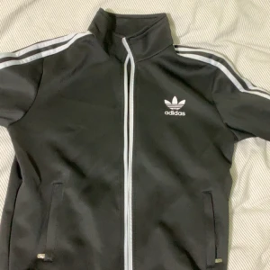 Svart Adidas track jacket med vita ränder - Svart Adidas track jacket med klassiska vita ränder längs ärmarna och vit dragkedja. Jackan har hög krage, två fickor med dragkedja och Adidas logga på bröstet. Perfekt för en sportig och chill stil.