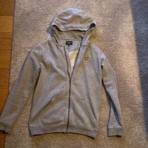 Grå hoodie från Lyle & Scott Junior - Snygg grå hoodie från Lyle & Scott Junior med dragkedja och huva. Klassisk gul logga på bröstet och två praktiska fickor framtill. Perfekt för chill dagar och enkel att matcha med jeans eller joggers.