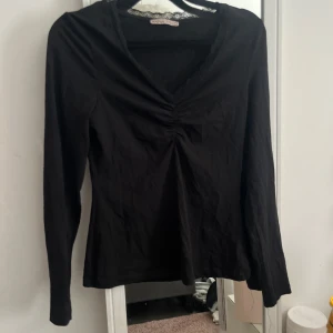 Svart långärmad topp med spets - Svart långärmad topp från Anna Field med snygg v-ringning och spetskant. Toppen har rynkade detaljer framtill som ger en cool touch. Mjuk bomullskänsla och figurnära passform, perfekt att styla med jeans eller kjol.