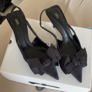 Svarta pumps med rosett från Zara - Snygga svarta pumps från Zara med spetsig tå och elegant rosett framtill. Skorna har smal klack och slingback-rem för en chic look. Materialet är matt syntet med tygrosett som ger extra stilpoäng. Perfekta för dig som gillar att sticka ut med detaljer. 