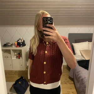Vinröd stickad kofta från H&M - Säljer en vinröd stickad kofta från H&M med korta ärmar och snygga guldfärgade knappar framtill. Koftan har ett klassiskt kabelstickat mönster och ribbade kanter. Perfekt att slänga över en t-shirt för en chill och trendig look. Kanske passar bättre storlek M