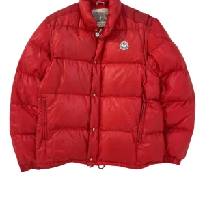 Röd Moncler jacka vintage - Säljer en fet röd dunjacka från Moncler med klassisk logga på bröstet. Jackan har hög krage, dragkedja och tryckknappar framtill samt två sidofickor med dragkedja. Materialet är glansig polyester och fyllningen är dun, perfekt för kalla dagar.