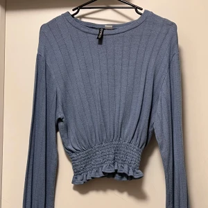 Blå ribbad tröja från Bershka - Säljer en blå långärmad tröja från Bershka med ribbad struktur och smockad midja som avslutas med en liten volangkant. Tröjan har rund hals och är i en mjuk, stretchig kvalitet som sitter snyggt på kroppen.