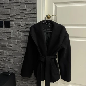 Svart omlottkappa från Vero Moda - Snygg svart kappa från Vero Moda med bred krage och omlottmodell. Jackan har ett matchande bälte i midjan och långa ärmar. Materialet är mjukt och känns som ull, perfekt för kyliga dagar. Stilren och enkel design som funkar till många outfits.