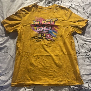 Gul Nike t-shirt med tryck - Gul t-shirt från Nike med färgstarkt 'Just Do It'-tryck i rosa, blått och svart på bröstet. Klassisk rund hals och korta ärmar. Tillverkad i 100% bomull för skön känsla. Perfekt för dig som gillar sportig och färgglad stil. Inga defekter. Priset kan diskuteras. 