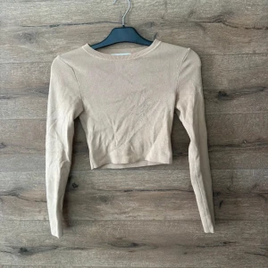 Beige croppad topp från Zara - Snygg croppad topp från Zara i beige färg. Modellen har lång ärm och rund halsringning med ribbad kant. Tunn och mjuk, perfekt att matcha med jeans eller kjol. Enkel och stilren design som funkar till många looks.