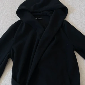 Svart omlott hoodiejacka med bälte - Svart hoodiejacka från NA-KD med omlott-design och bälte i midjan. Jackan har långa ärmar och en stor huva som ger en chill vibe. Perfekt för dig som gillar enkel och stilren look. Materialet är mjukt och känns skönt mot huden. Helt oanvänd ! 