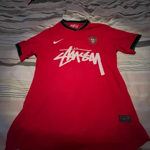 Portugal x Stüssy röd fotbollströja Nike M - Säljer en röd Portugal fotbollströja i samarbete med Stüssy från Nike. Tröjan har korta ärmar med gröna detaljer vid krage och ärmslut, samt Portugal-emblem på bröstet och stor Stüssy-logga framtill. Tillverkad i Dri-Fit material som andas.
