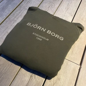 Olivgrön Björn Borg hoodie med huva - Snygg olivgrön hoodie från Björn Borg med tryck 'BJÖRN BORG STOCKHOLM 1956' på bröstet. Tröjan har huva med snörningshål, långa ärmar och en klassisk känguruficka framtill. Perfekt för chill eller häng med kompisar.