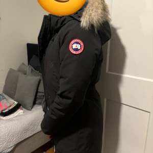Svart Canada Goose dunparkas med päls - Säljer en svart Canada Goose parkas med ikonisk patch på ärmen och fluffig päls runt huvan. Jackan är lång, har dragkedja och knappar framtill, samt en snygg och varm design som är perfekt för kalla dagar. Flyttar utomlands så har tyvärr ingen användning för den längre, köpt på NK i Göteborg och kvittot är borttappat men garanterar att den är äkta. Storlek XS, lite slitage på ärmarna, det går även att tvätta den på kemtvätt med svart färg som gör att den ser intill ny ut. Använt 3 vintrar. 