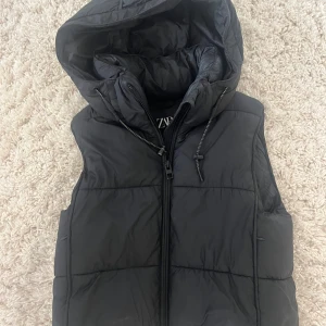 Svart dunväst med huva från Zara - Snygg svart dunväst från Zara med stor vadderad huva och dragkedja framtill. 