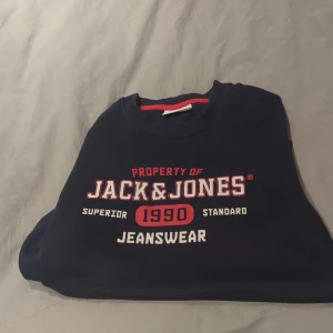 Mörkblå sweatshirt från Jack & Jones - Snygg mörkblå sweatshirt från Jack & Jones med tryck i vitt och rött på bröstet. Tröjan har rund hals, långa ärmar och ribbade muddar. Perfekt för dig som gillar streetwear och vill ha en casual look. Materialet är mjuk bomull som känns skönt mot huden.