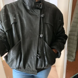 Svart retro 80-tals skinnjacka från Faris - Säljer en svart skinnjacka från Faris, från 80-talet, med coola detaljer som höga kragen med spänne, stora fickor och snygga knappar framtill. Jackan har en loose passform och är helfodrad med Faris-logga på insidan. Väldigt skön och varm. 