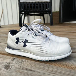 Vita golfskor från Under Armour - Snygga vita golfskor från Under Armour med marinblå detaljer och logga på sidan. Skorna har perforerat mönster för extra ventilation, snörning och en platt sula. Ovandelen är i syntetmaterial och insidan är vadderad för skön komfort. Bra grepp under för just golf. Finns små slitningar men absolut inget betydande. 