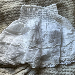 Vit broderad kortkjol från H&M - Supersöt vit kortkjol från H&M med bred smock i midjan och broderade detaljer. Kjolen har volanger och spetskant, vilket ger en romantisk och fräsch vibe. Perfekt för varma dagar och lätt att matcha med en enkel topp. Bandet i midjan är lite trasigt men syns knappt.