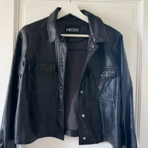 Svart skinnjacka från Pieces - Snygg svart skinnjacka från Pieces med klassisk krage och två stora bröstfickor med knappar. Jackan har långa ärmar och tryckknappar framtill. Perfekt för dig som vill ha en cool och stilren look.