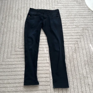 Svarta Replay Anbass jeans straight fit - Säljer ett par svarta Replay Anbass jeans med straight fit och klassisk femficksdesign. Jeansen har Replay-logga vid fickan, knappgylf och bälteshällor. Materialet är mjukt och stretchigt jeans, perfekt för dig som vill ha en clean och stilren look.