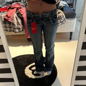 Spicy bootcut jeans med detaljer - Säljer ett par snygga spicy jeans med låg midja och bootcut passform. De är blå med slitningar och patchade detaljer på benen, har flera fickor och dubbla knappar i midjan. Jeansen är gjorda i klassiskt denim. Midjemått på bilder, aldrig använda då dom är för små för mig. Prislapp kvar